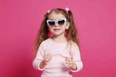 Pembe arka planda güneş gözlüklü, gülümseyen küçük kız.