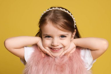 Sarı arka planda, saç bandı ve kürklü yelek giymiş gülümseyen küçük bir kızın portresi.