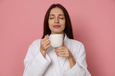 Pembe arka planda bir fincan kahveli bornoz giymiş bir kadın.