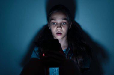 Siber tehlike. Karanlık arka planda telefon kullanan korkmuş bir kız.