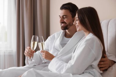 Yataklarında köpüklü şarap bardakları olan bornozlu mutlu bir çift.