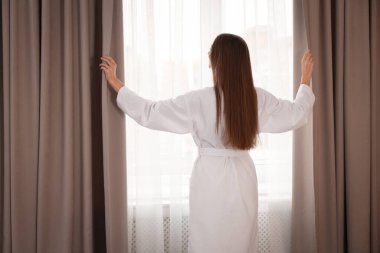 Beyaz bornozlu kadın perdeleri açıyor.