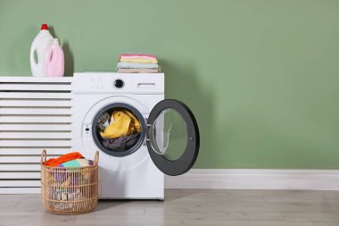 Modern çamaşır makinesi, deterjanlar ve açık yeşil duvarın yanındaki kıyafetler.