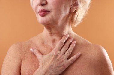 Yaşlı kadın soluk turuncu arka planda boynuna dokunuyor, yakın plan.