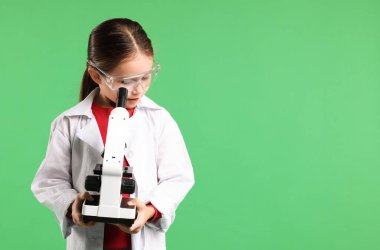 Laboratuvar önlüklü, koruyucu gözlüklü yeşil arka planda mikroskobu olan küçük bir kız. Mesaj için yeri var. Çocuk ve bilim