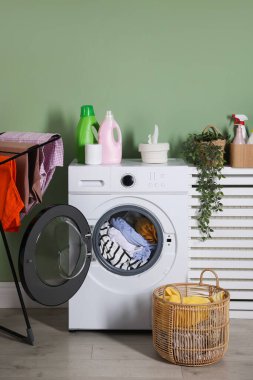 Modern çamaşır makinesi, deterjanlar, ev bitkisi ve açık yeşil duvarın yanındaki kıyafetler.