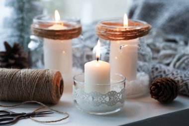 DIY Noel fenerleri. Pencere pervazında cam kavanozlarda, konilerde, sicimde ve makaslarda yanan mumlar, yakın plan.