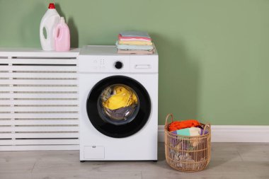 Modern çamaşır makinesi, deterjanlar ve açık yeşil duvarın yanındaki kıyafetler.
