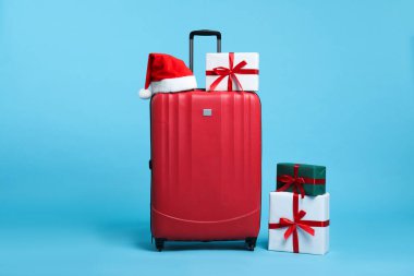 Noel tatili. Kırmızı bavul, Noel Baba şapkası ve açık mavi arka planda hediye kutuları.