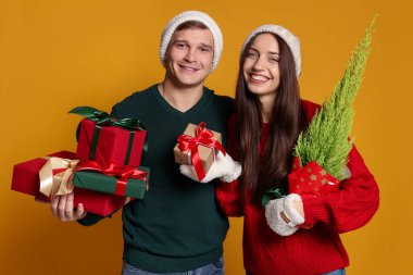 Şapkalı mutlu bir çift, sarı arka planda bir Noel ağacı ve hediyeleri.