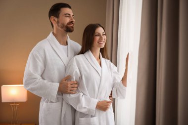 Otel odasının penceresinin yanında bornoz giyen mutlu çift.