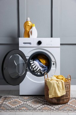 Modern çamaşır makinesi, deterjanlar ve gri duvarın yanındaki kıyafetler.