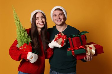 Şapkalı mutlu bir çift, sarı arka planda bir Noel ağacı ve hediyeleri.