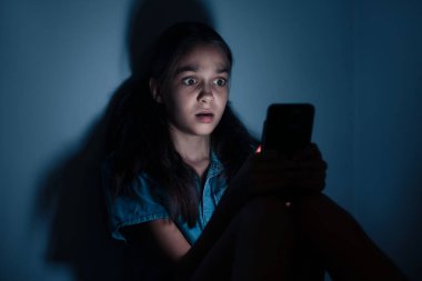 Siber tehlike. Karanlık arka planda telefon kullanan korkmuş bir kız.