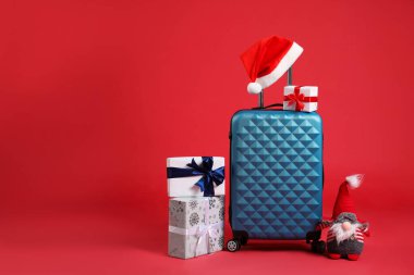 Noel tatili. Şık bavul, Noel Baba şapkası, hediye kutuları ve kırmızı arka planda dekoratif cüce. Metin için boşluk