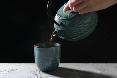 Gri masadaki çaydanlıktan fincana sıcak çay dolduran kadın, yakın plan.