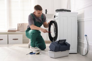 Üniformalı tamirci evde tornavidayla bozuk çamaşır makinesini tamir ediyor.