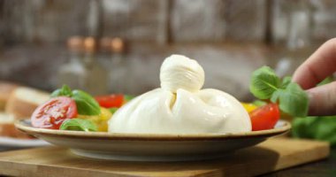 Ahşap masada nefis burrata peyniri ve domateslere fesleğen koyan bir kadın.