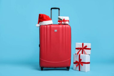 Noel tatili. Kırmızı bavul, Noel Baba şapkası ve açık mavi arka planda hediye kutuları.