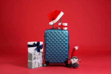 Noel tatili. Şık bavul, Noel Baba şapkası, hediye kutuları ve kırmızı arka planda dekoratif cüce.