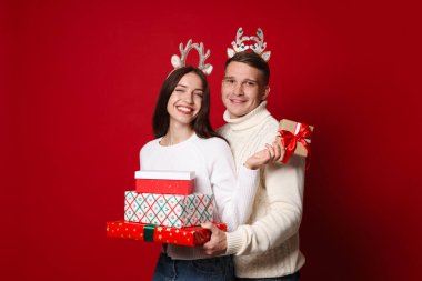 Kırmızı arka planda Noel hediyeleri olan ren geyiği bantlı mutlu çift