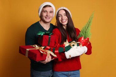 Şapkalı mutlu bir çift, sarı arka planda bir Noel ağacı ve hediyeleri.