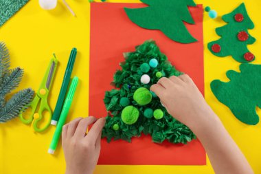 Küçük çocuk sarı masada Noel kartpostalı hazırlıyor. Çocuk el yapımı.