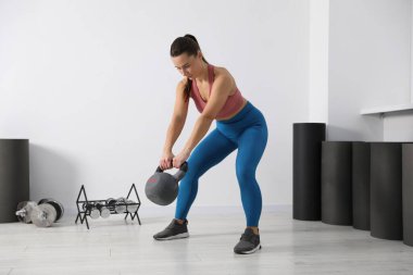 Spor salonunda beyaz duvarın yanında kettlebell ile egzersiz yapan atletik bir kadın.