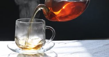 Gri masadaki çaydanlıktan fincana sıcak çay dolduran kadın, yakın plan.