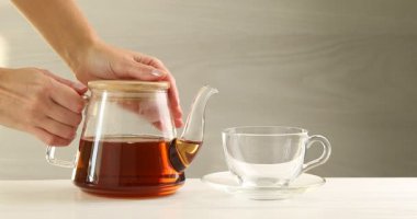 Beyaz ahşap masada çaydanlıktan fincana sıcak çay dolduran kadın, yakın plan.