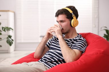 Kulaklıklı genç adam evde kahve poşeti koltukta müzik dinliyor.