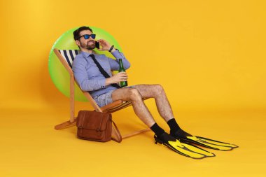 Elinde içki şişesi olan bir iş adamı güverte koltuğunda turuncu arka planda akıllı telefonla konuşuyor.