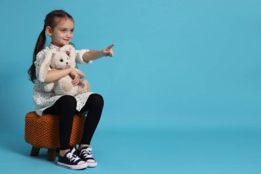 Oyuncak tavşanı olan sevimli küçük bir kız puf üzerinde oturuyor ve açık mavi arka planda bir şeye işaret ediyor. Metin için boşluk