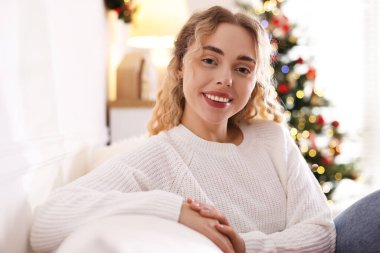 Evdeki bulanık ışıklara karşı kanepede süveter giymiş gülümseyen kadın portresi. Mutlu Noeller ve mutlu yıllar.