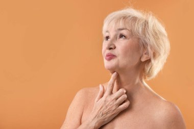 Yaşlı kadın soluk turuncu arka planda boynuna dokunuyor. Metin için boşluk