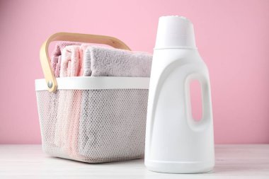 Çamaşır sepetinde havlular ve bir şişe deterjan beyaz ahşap masada pembe arka planda, yakın plan.