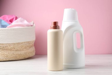 Çamaşır sepetinde elbiseler ve beyaz ahşap masada pembe arka planda bir şişe deterjan var.