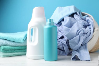 Çamaşır sepeti, beyaz ahşap masada açık mavi arka planda giysi ve şişe deterjan.