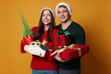 Şapkalı mutlu bir çift, sarı arka planda bir Noel ağacı ve hediyeleri.