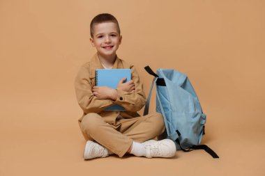 Bej arka planda not defteri ve sırt çantası olan sevimli küçük bir çocuk.