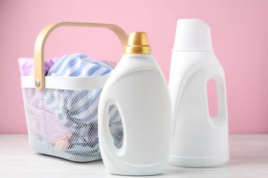 Çamaşır sepetinde giysiler ve beyaz ahşap masa üzerindeki deterjan pembe arka planda, yakın plan.