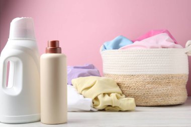 Çamaşır sepetinde elbiseler ve beyaz ahşap masada pembe arka planda bir şişe deterjan var.