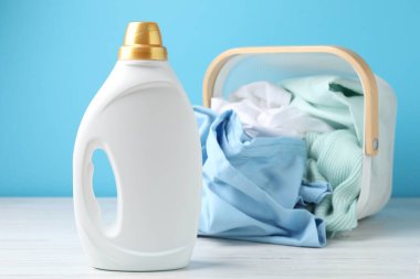 Çamaşır sepetinde elbiseler ve beyaz ahşap masada bir şişe deterjan açık mavi arka planda, yakın plan