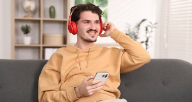 Kulaklıklı, akıllı telefonlu mutlu bir adam evdeki kanepede müzik dinliyor.