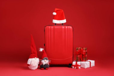 Noel tatili. Şık bavul, Noel Baba şapkası, hediye kutuları ve kırmızı arka planda dekoratif cüceler.