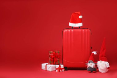 Noel tatili. Şık bavul, Noel Baba şapkası, hediye kutuları ve kırmızı arka planda dekoratif cüceler. Metin için boşluk