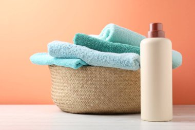 Çamaşır sepetinde havlular ve beyaz ahşap masada mercan arka planda bir şişe deterjan var.