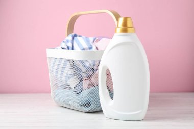 Çamaşır sepetinde elbiseler ve beyaz ahşap masada pembe arka planda bir şişe deterjan.