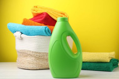 Çamaşır sepetinde havlular ve beyaz ahşap masada bir şişe deterjan, sarı arka planda, yakın plan.