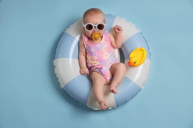 Güneş gözlüklü, oyuncak ördekli, şişme yüzüklü, açık mavi arka planda şirin bir bebek.